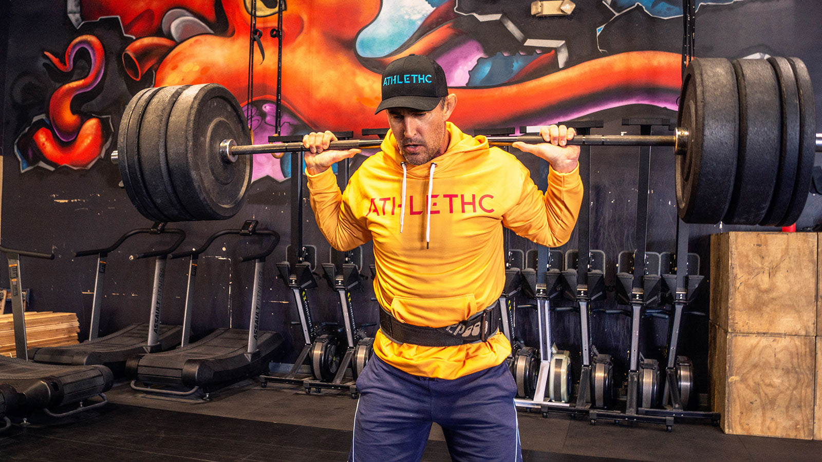 Man lifting weights in a gym with colorful wall art in the background