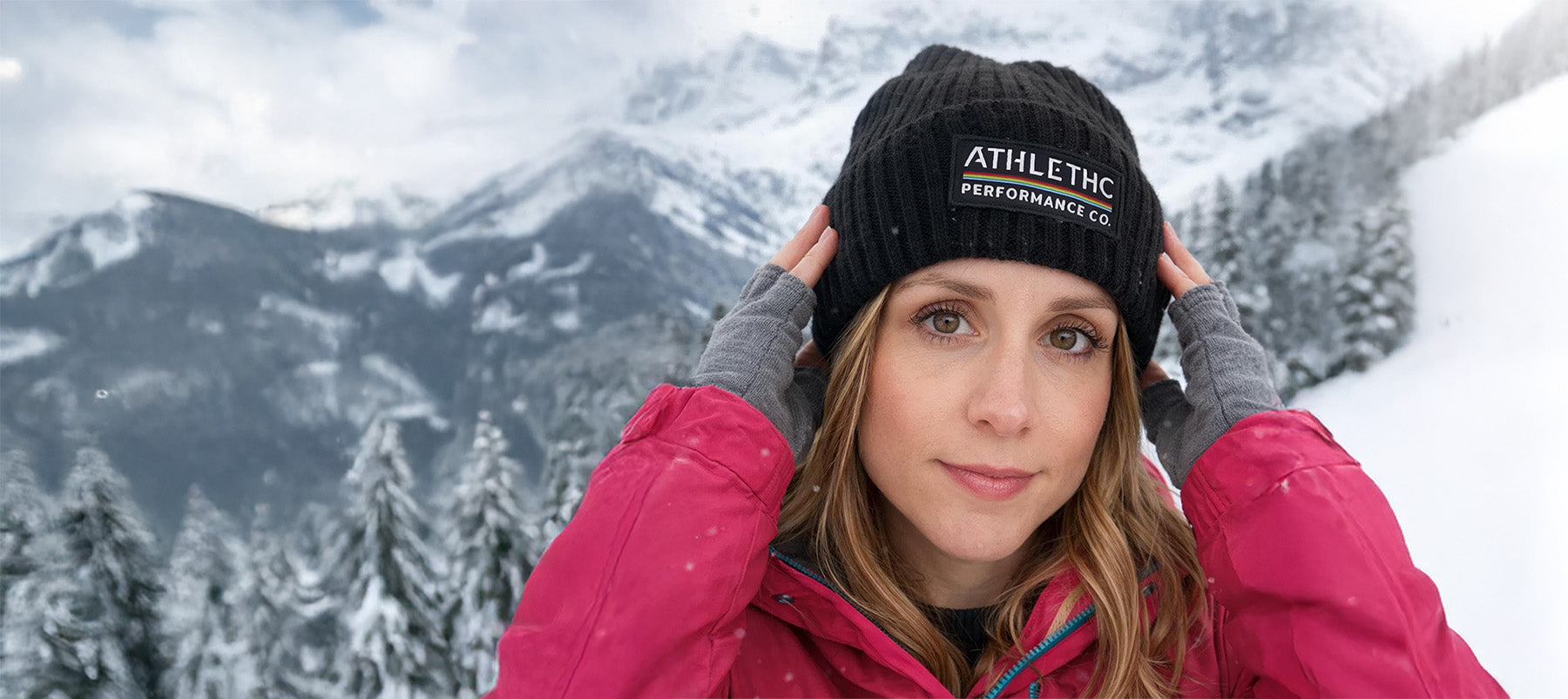 Woman wearing a black beanie with 'ATHLETHC Performance Co' text in a snowy mountain setting