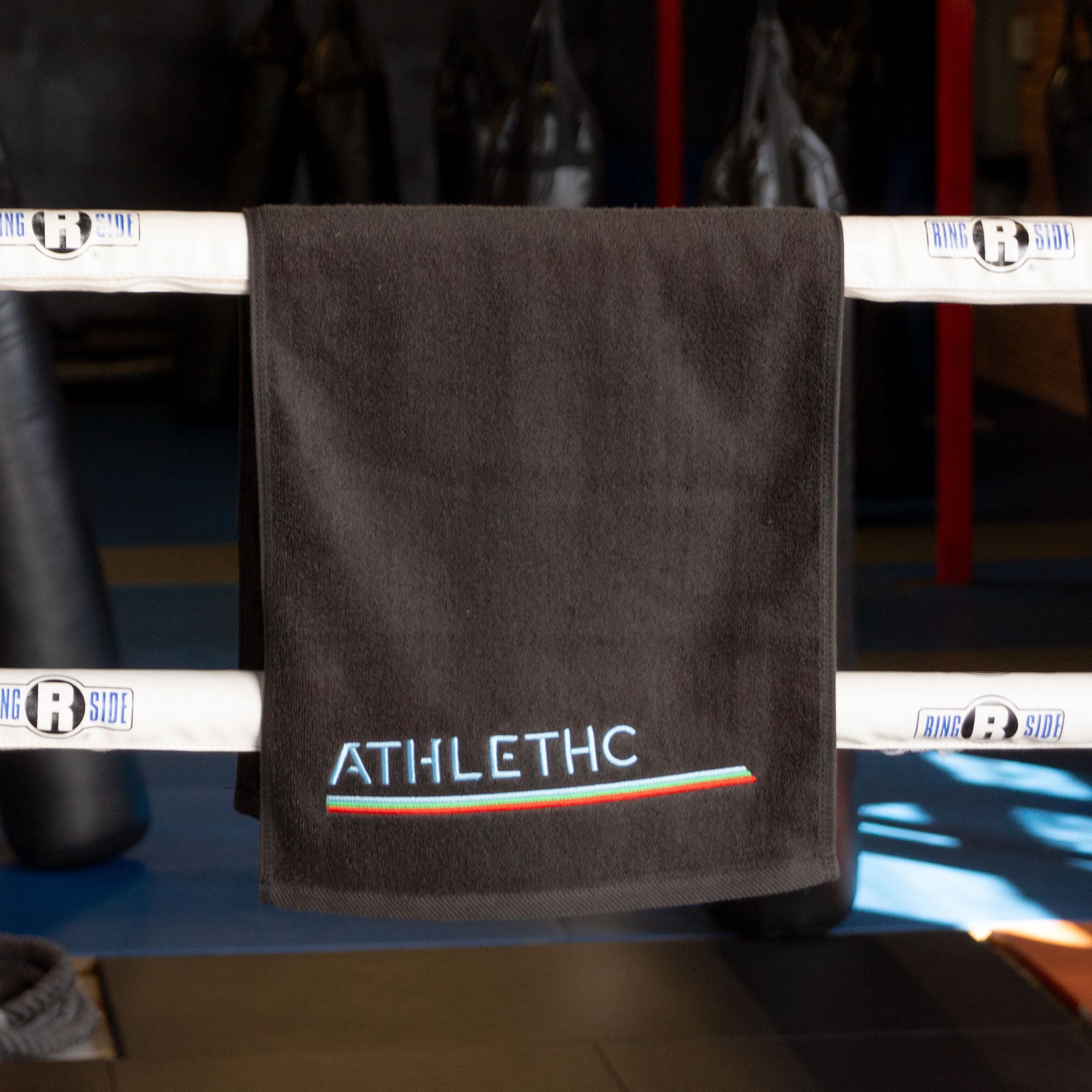 A black cotton gym towel with the word 'ATHLETHC' embroidered on it, hanging on a boxing ring.