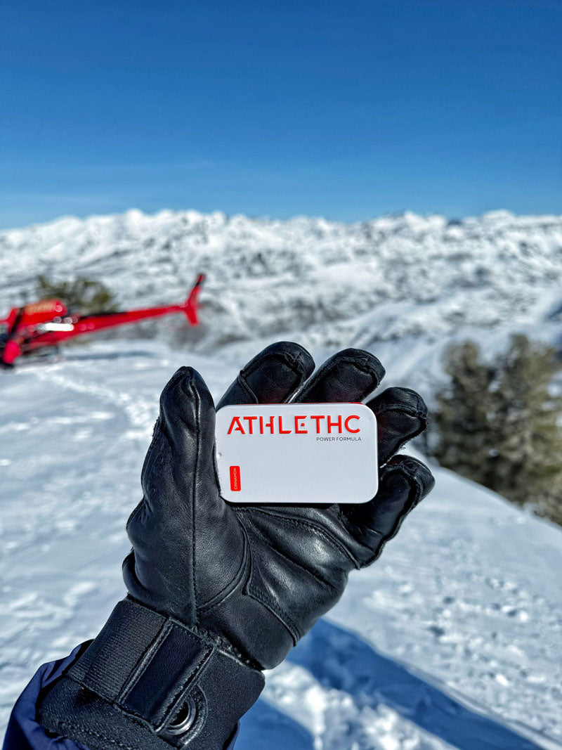 Skier on a mountain with a helicopter holding a tin of ATHLETHC Power Microdose THC Performance mints.