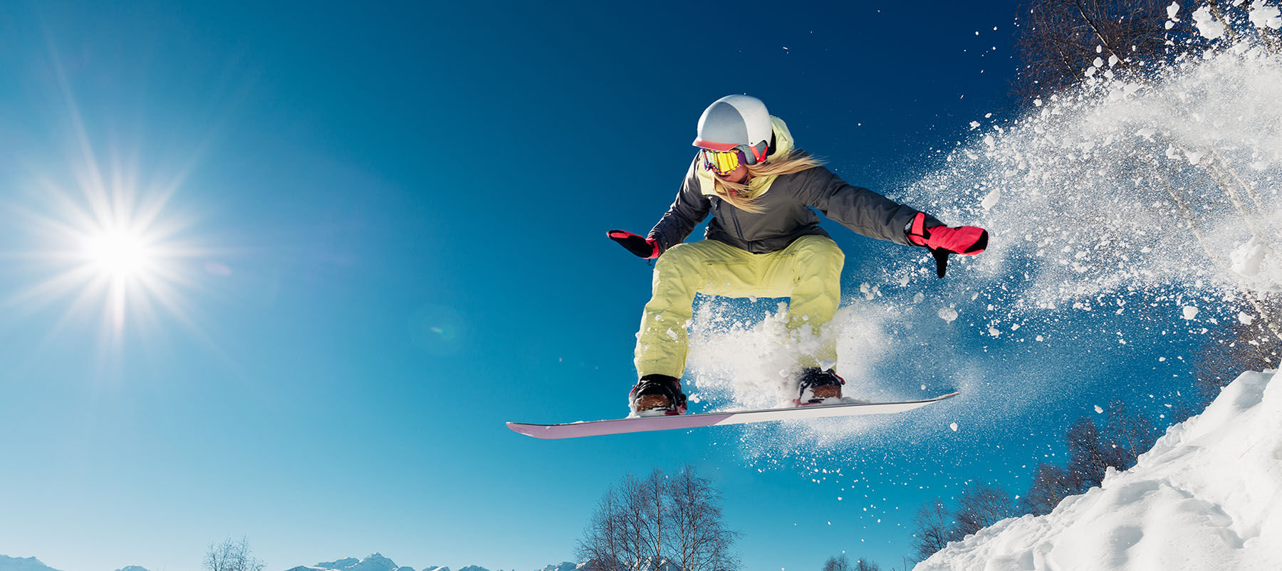 Person snowboarding in mid-air against a blue sky with sun and snow.