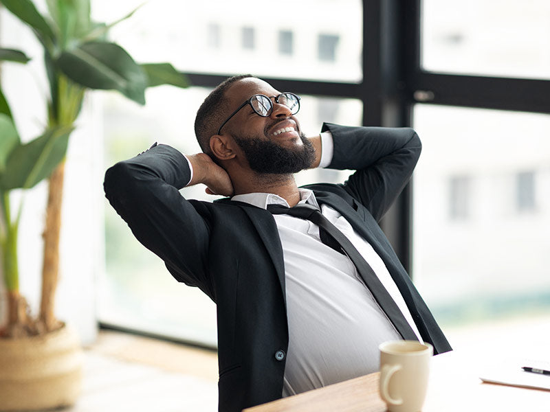 Man in formal attire with a beard and glasses, sitting at a desk with a cup of coffee, smiling.