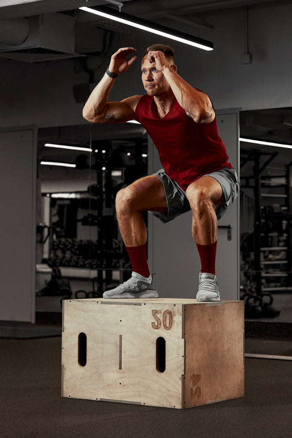 Man performing a box jump on a wooden box in a gym setting.