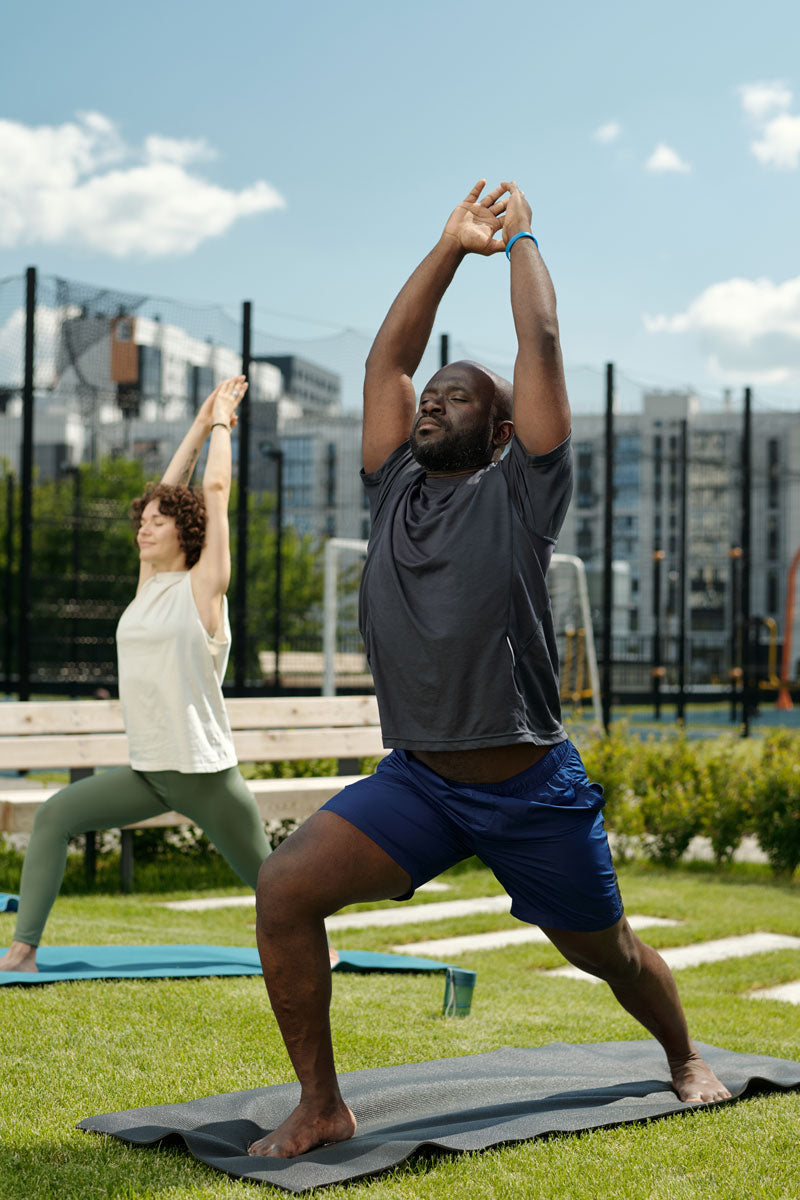 Two people practicing yoga outdoors on a grassy area with buildings in the background.